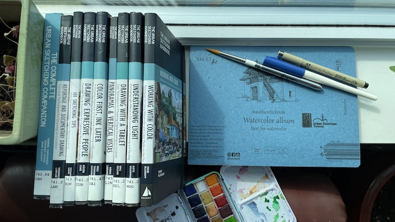 a stack of Urban Sketchers handbooks, with a sketchbook, watercolour palette and assortment of pens and brushes